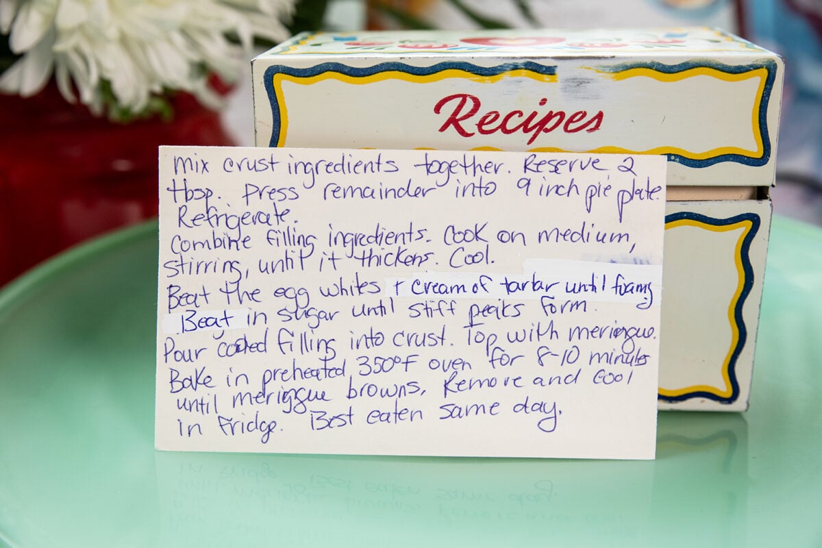 A handwritten recipe card in front of a colorful recipe box, with flowers blurred in the background. The card lists steps for making a pie, including preparing crust, filling, and meringue topping.