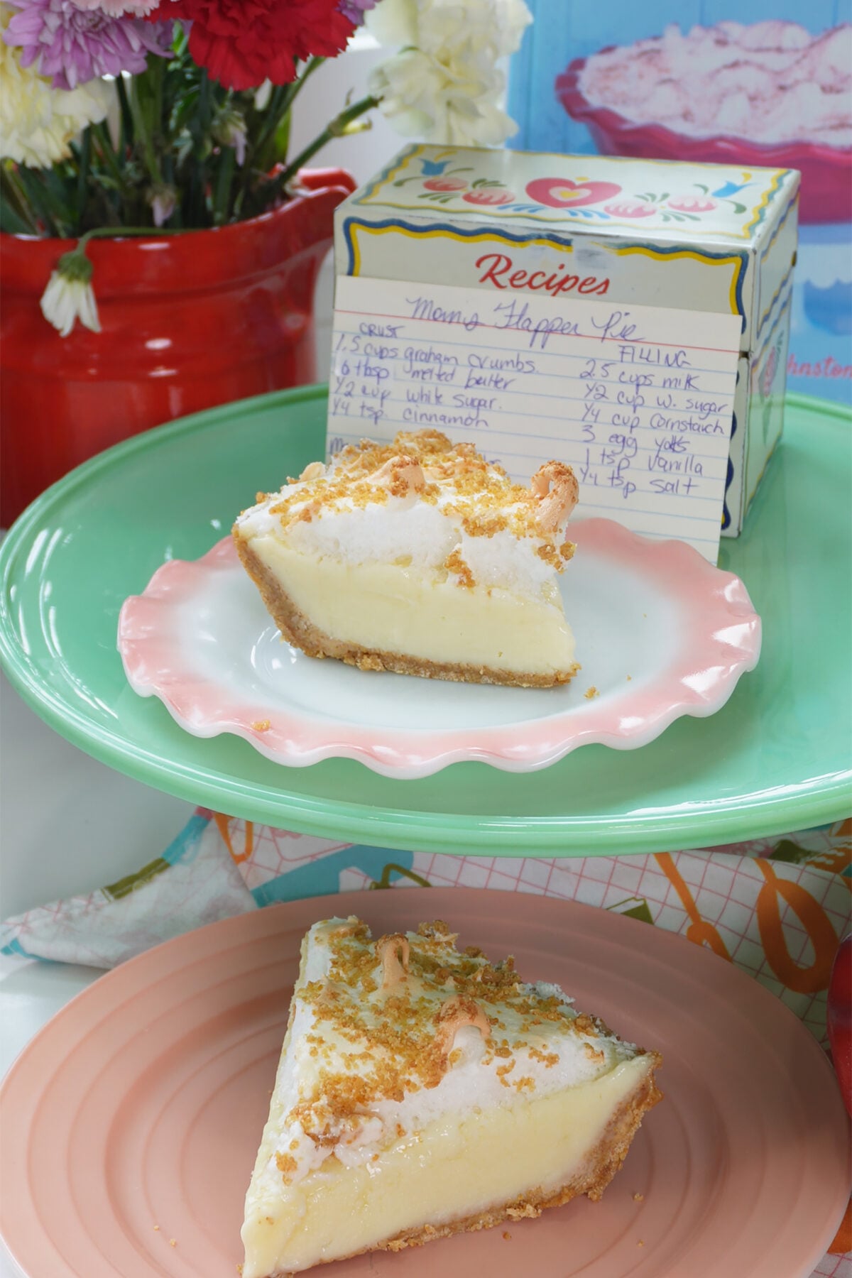 A slice of creamy pie with a crumb crust sits on a pink plate, with another slice on a mint-green stand. Behind them is a floral bouquet and a recipe box with a handwritten recipe card visible.