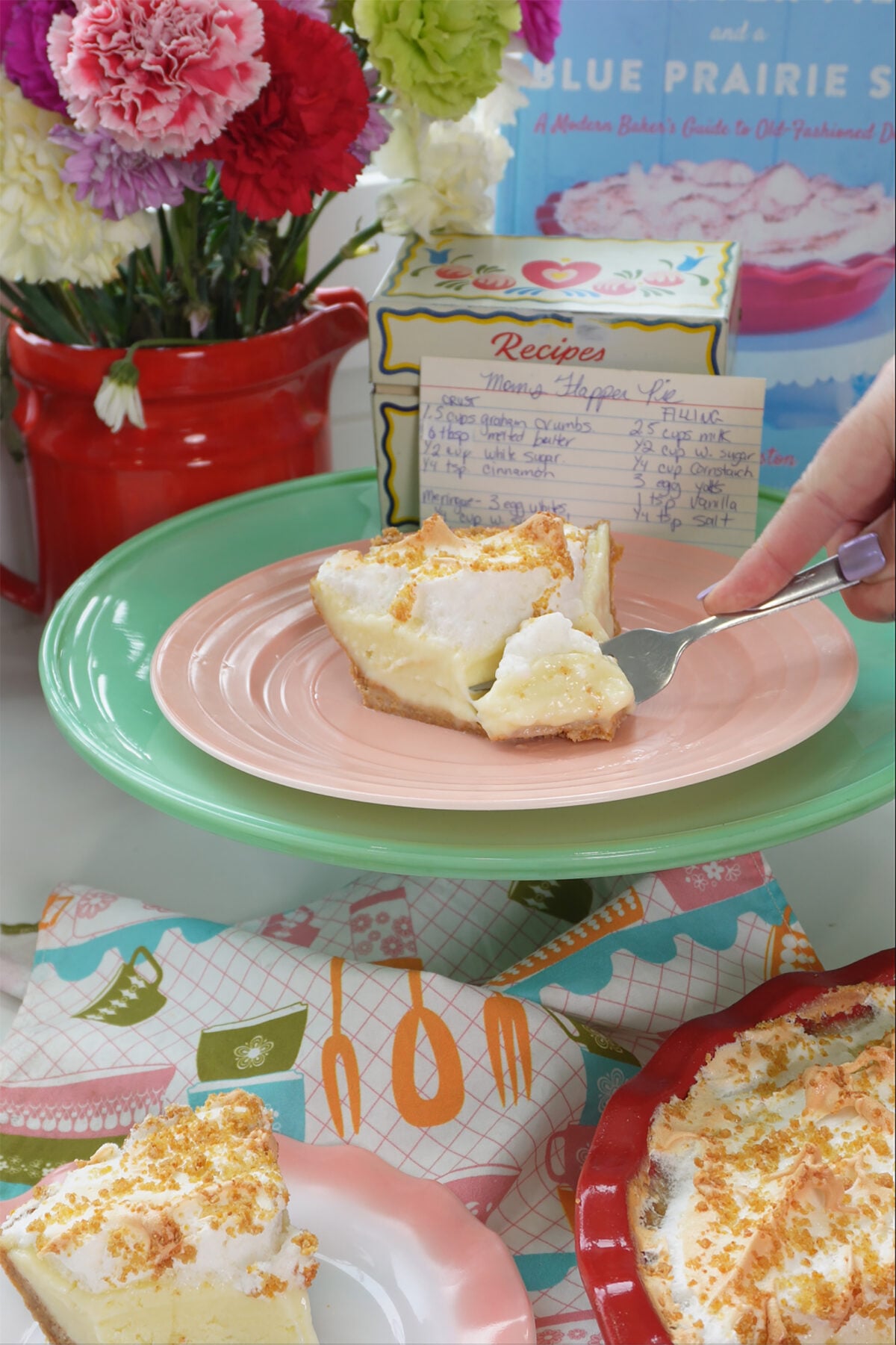 A slice of meringue-topped pie on a pink plate, with a fork lifting a bite, colorful flowers, a handwritten recipe card, and a pie dish on a patterned napkin in the background.