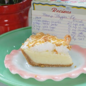 A slice of cream pie with a crumb crust and meringue sits on a pink plate. Behind it is a recipe box with a handwritten recipe card and a red pot with green leafy plants.