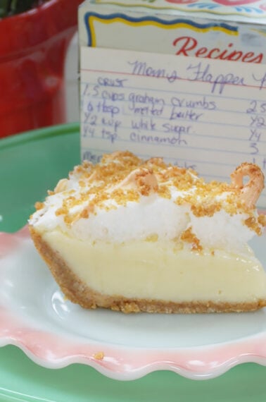 A slice of cream pie with a crumb crust and meringue sits on a pink plate. Behind it is a recipe box with a handwritten recipe card and a red pot with green leafy plants.