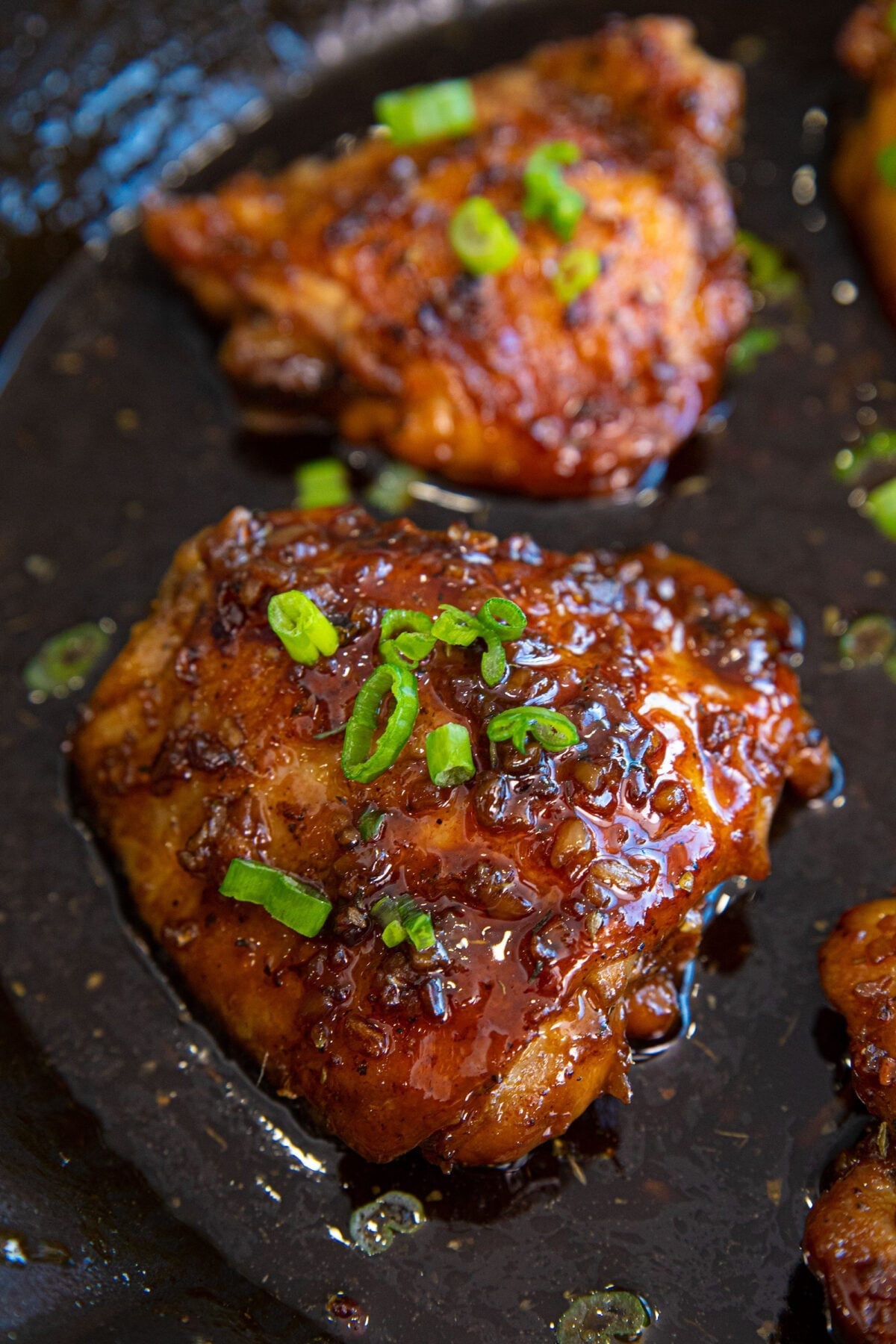 Glazed chicken thigh garnished with chopped green onions on a dark surface, showing a shiny, caramelized coating and visible seasoning.