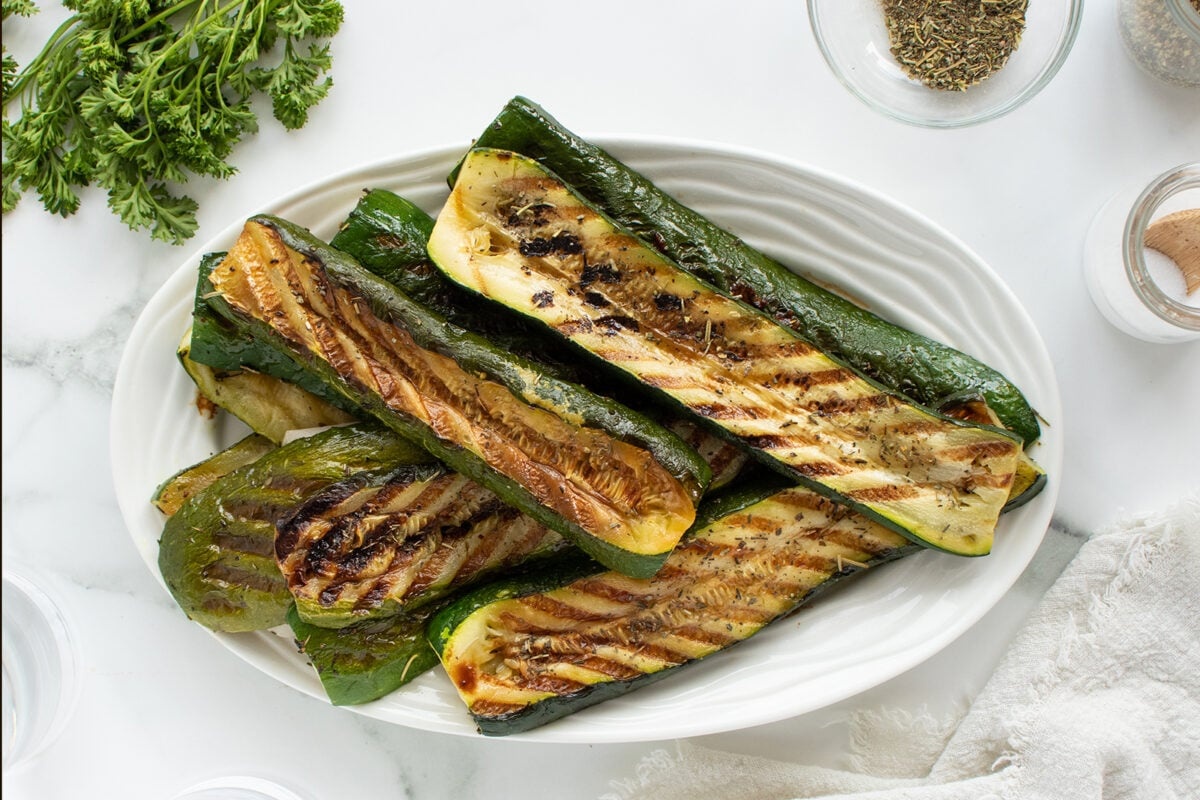 Grilled zucchini halves with char marks are arranged on a white oval plate. Fresh parsley, spices, and a cloth napkin are visible around the plate on a white surface.