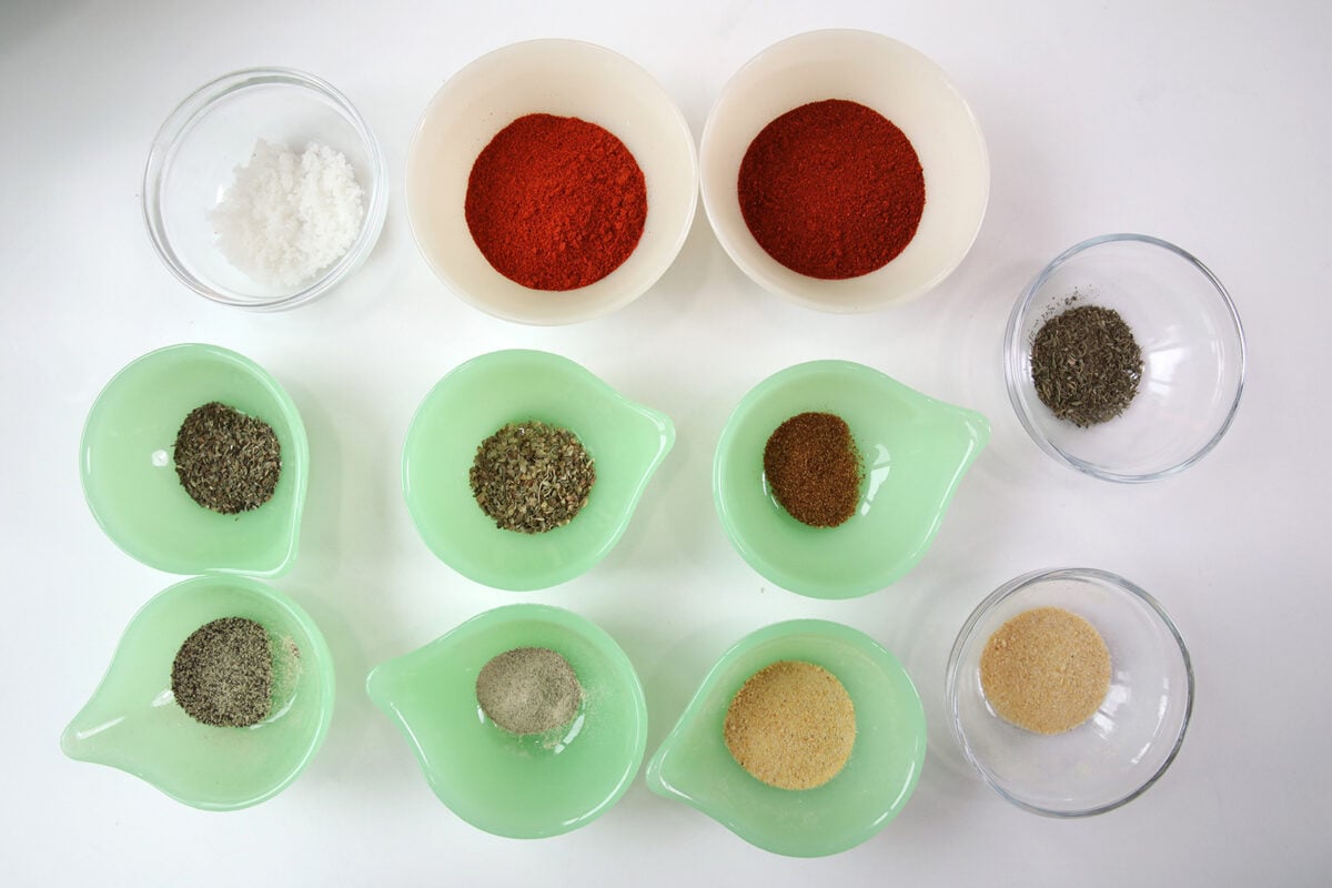 Nine small bowls with various dry spices, including red powders, salt, greenish herbs, and beige granules, are arranged neatly on a white surface.