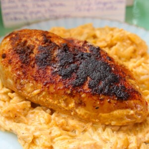 A seasoned, blackened chicken breast sits on a bed of creamy, cheesy pasta on a white plate. A handwritten recipe card is blurred in the background.