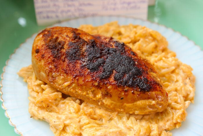 A seasoned, blackened chicken breast sits on a bed of creamy, cheesy pasta on a white plate. A handwritten recipe card is blurred in the background.