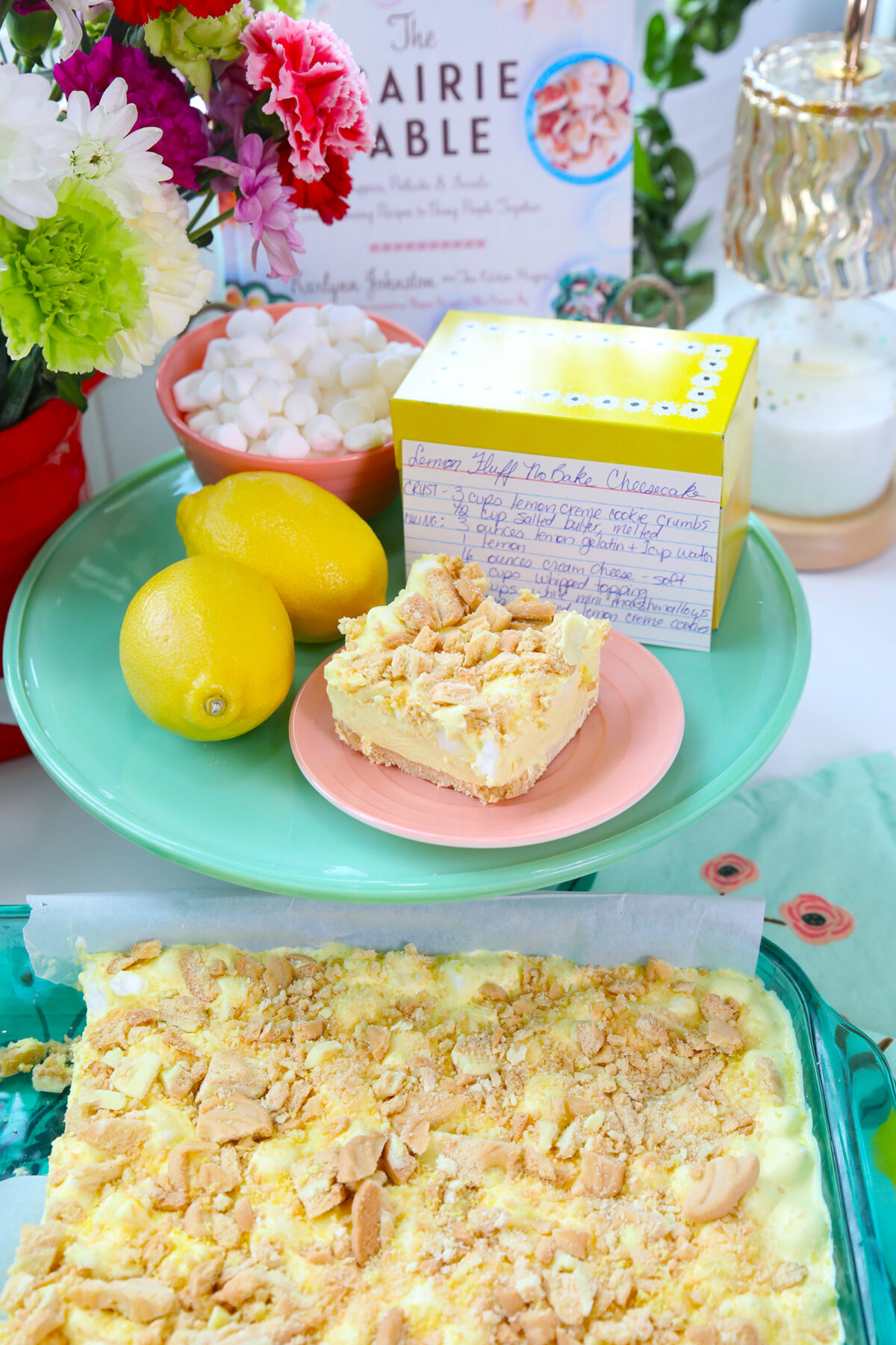 Lemon-fluff cheesecake bars on a plate, with lemons, a recipe card, marshmallows, and flowers beside a tray of the dessert. Cookbooks and candles appear in the bright, colorful background.