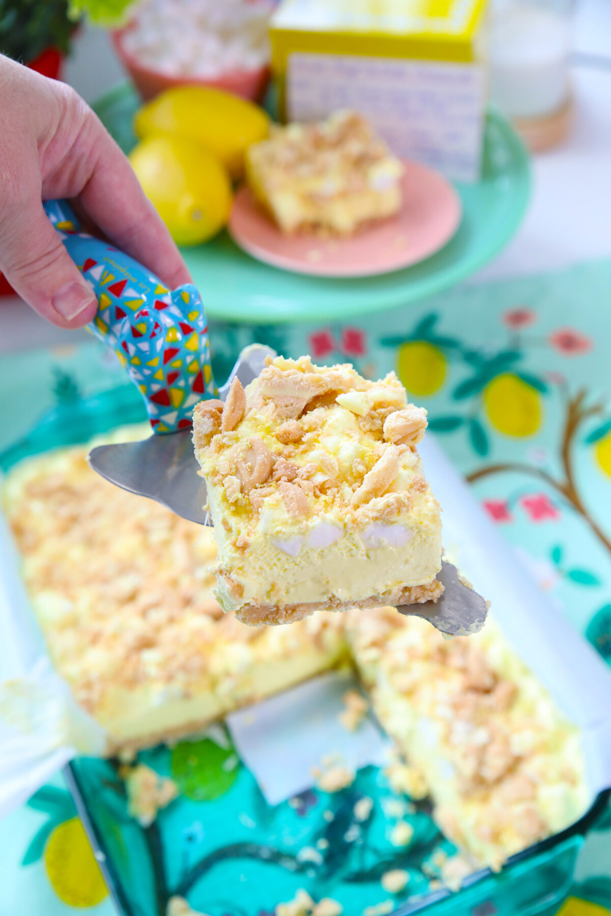 A hand holds a colorful spatula lifting a lemon dessert bar with a crumbly topping from a glass baking dish. Lemons and a box of pudding mix are visible in the background.