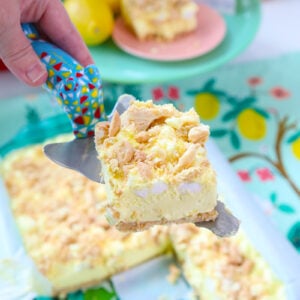A hand holds a colorful spatula lifting a lemon dessert bar with a crumbly topping from a glass baking dish. Lemons and a box of pudding mix are visible in the background.