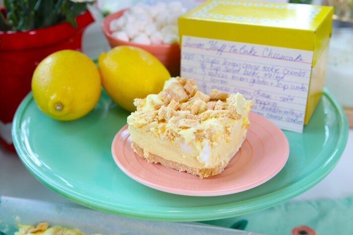 A slice of lemon cheesecake topped with crumbs sits on a pink plate. Nearby are fresh lemons, a yellow recipe box with a handwritten recipe, and a bowl of mini marshmallows, all arranged on a green platter.