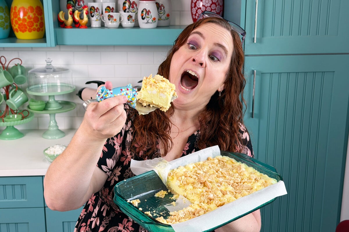 A woman with long brown hair excitedly holds a tray of dessert bars in one hand and a large serving in the other, about to take a big bite in a colorful kitchen.
