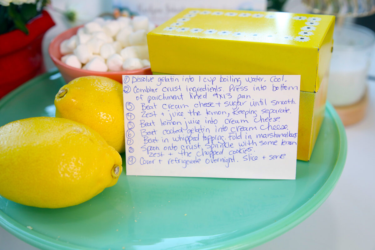 A handwritten recipe card next to two lemons and a yellow box, with a plate of marshmallows and a glass of milk blurred in the background. The recipe lists steps for a dessert, starting with dissolving gelatin.