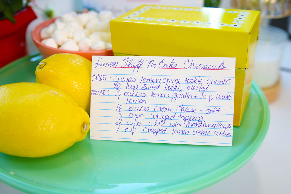 A recipe card for Lemon Fluff No Bake Cheesecake sits on a turquoise plate, next to two lemons and a yellow recipe box. Marshmallows are blurred in the background.