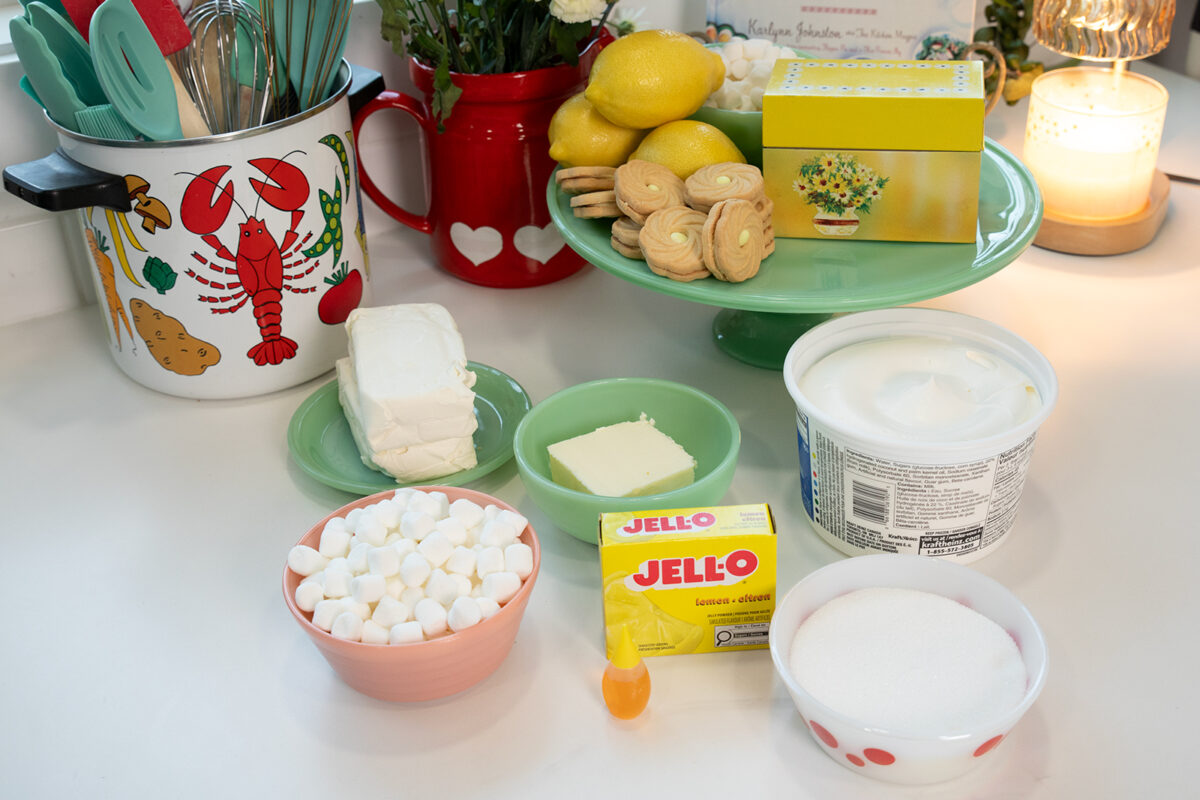 Various dessert ingredients on a kitchen counter, including marshmallows, cream cheese, butter, sugar, Jell-O, cookies, lemons, whipped topping, and utensils in a lobster-themed container.