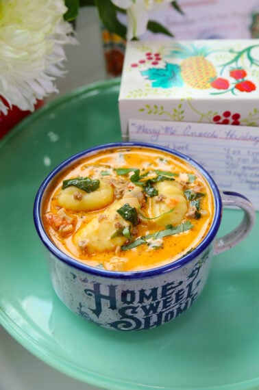 A mug labeled Home Sweet Home filled with creamy soup and dumplings, garnished with herbs, sits on a green plate. A recipe card and decorated box are in the background.