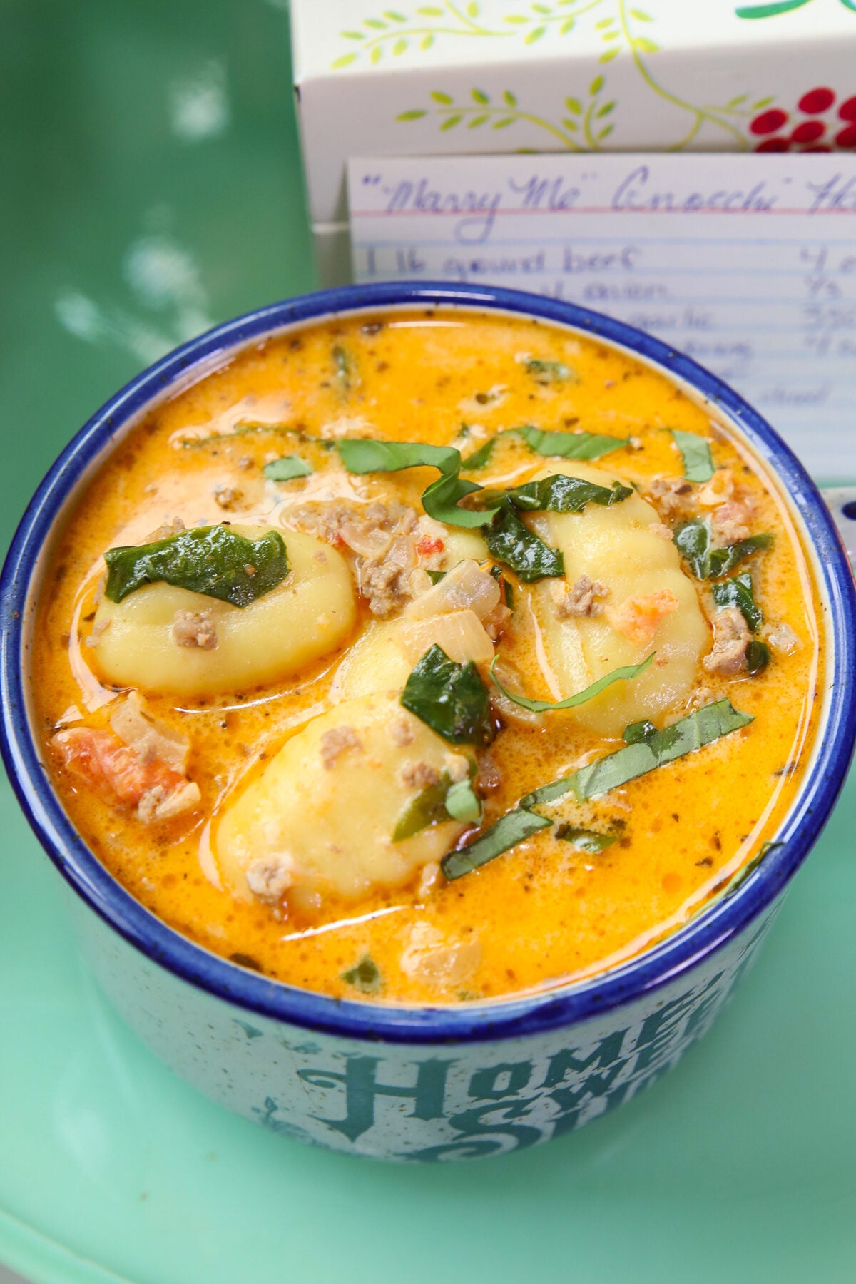 Creamy soup with gnocchi, ground meat, tomatoes, and spinach in a blue and white mug. A handwritten recipe card is slightly visible in the background.