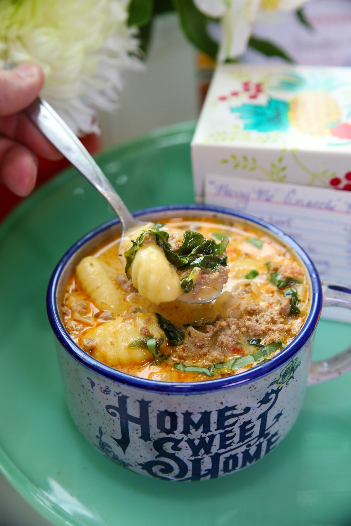 A hand holds a spoonful of creamy soup with gnocchi and greens above a blue Home Sweet Home mug filled with soup, set on a green plate. A floral recipe box is blurred in the background.
