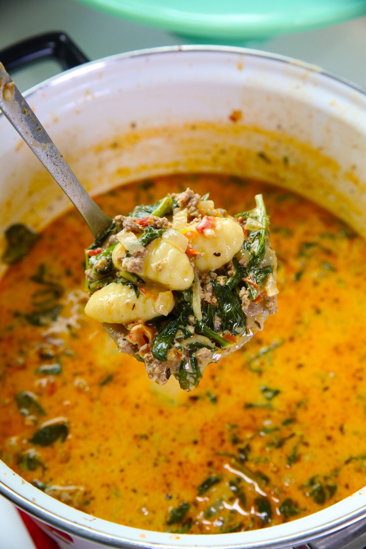 A close-up of a ladle lifting gnocchi, ground meat, and greens from a pot of creamy orange soup with herbs.