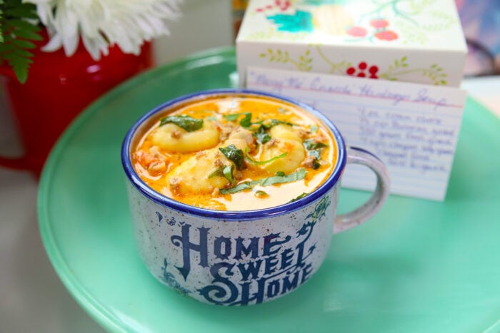 A mug labeled Home Sweet Home is filled with a creamy soup containing tortellini, spinach, and herbs. The mug sits on a green tray next to a floral card and a red container with white flowers.