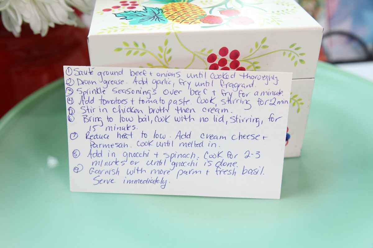 Handwritten recipe instructions on white paper, placed in front of a decorated recipe box with fruit illustrations. The paper lists cooking steps in blue ink, and both are on a green plate with a blurry background.