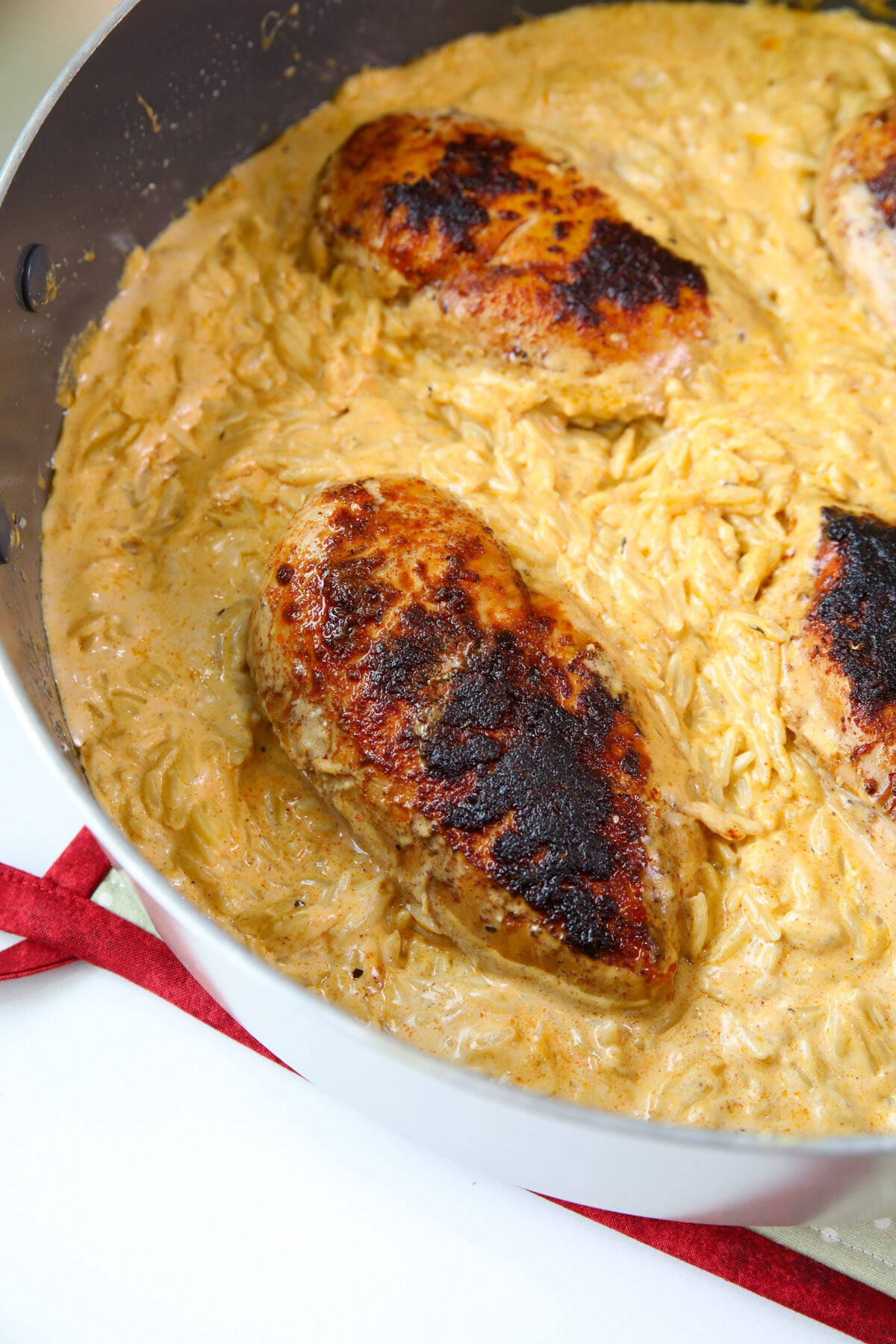Three seared chicken breasts rest in a creamy, golden sauce with orzo pasta inside a large skillet, shown from above on a white surface with a red cloth partially visible.