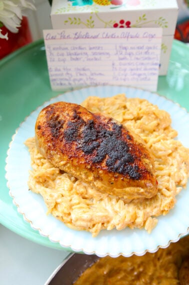 A blackened chicken breast rests on a bed of creamy orzo on a light blue scalloped plate. A recipe card for One Pan Blackened Chicken Alfredo Orzo is displayed in the background.