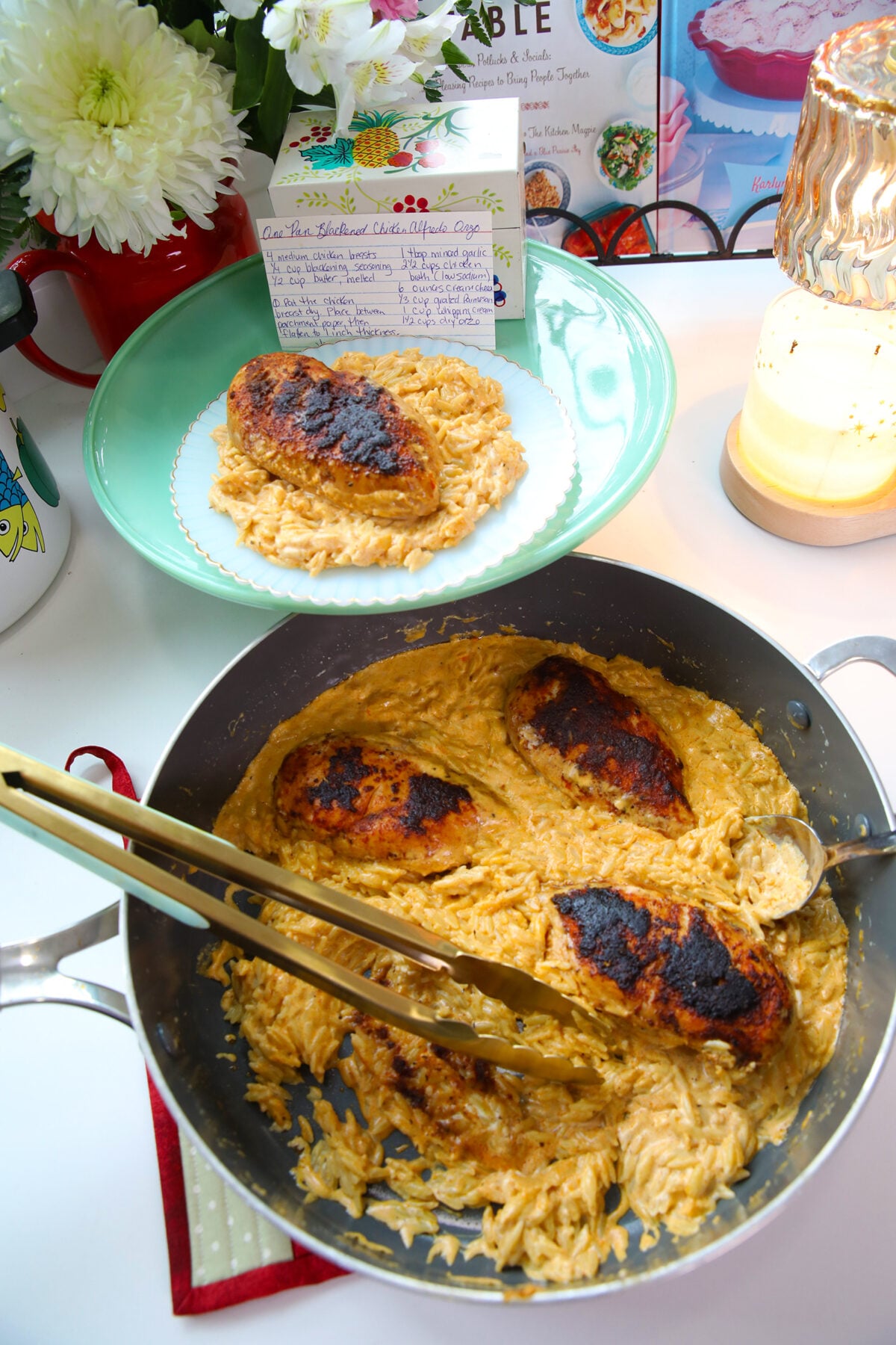 A skillet of creamy, blackened chicken Alfredo orzo sits in the foreground with gold tongs. Behind it, a single serving is plated on a light blue dish next to a printed recipe card, a vase of flowers, and a glowing candle.