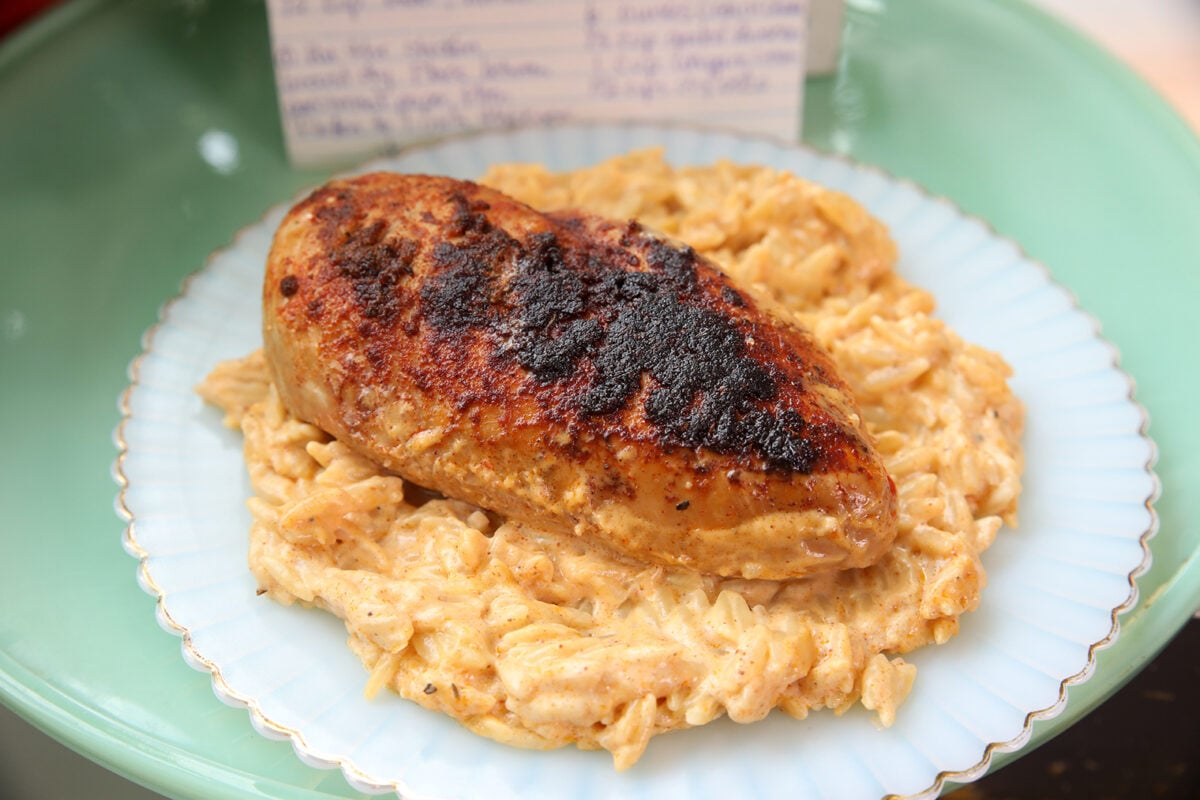 A piece of blackened, seasoned chicken breast sits on a mound of creamy, cheesy orzo pasta on a white plate, with a blurred recipe card in the background.