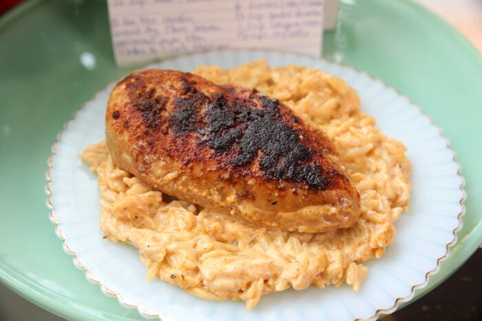 A piece of blackened, seasoned chicken breast sits on a mound of creamy, cheesy orzo pasta on a white plate, with a blurred recipe card in the background.
