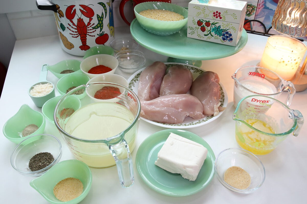 Various ingredients for a recipe are arranged on a counter, including raw chicken breasts, measuring cups with liquids, spices in small bowls, a block of cream cheese, butter, and grains on green and clear dishware.