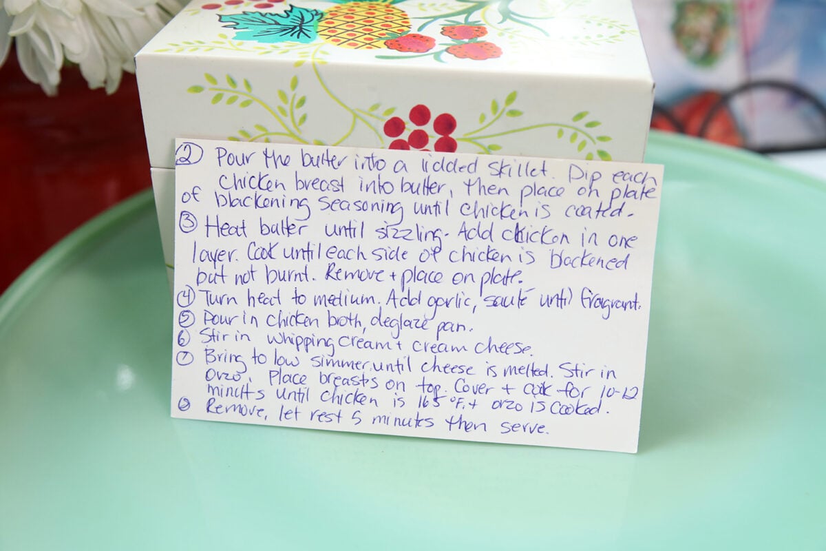 A handwritten recipe on a card next to a floral box, detailing steps for cooking chicken with butter, blackening seasoning, and cheese in a skillet. The card is propped up on a reflective surface.