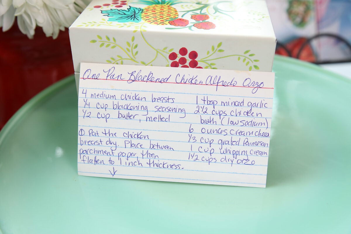 A handwritten recipe card for One Pan Blackened Chicken Alfredo Orzo sits in front of a decorated box on a light green plate. The card lists ingredients like chicken, blackened seasoning, butter, garlic, broth, cream cheese, parmesan, and orzo.