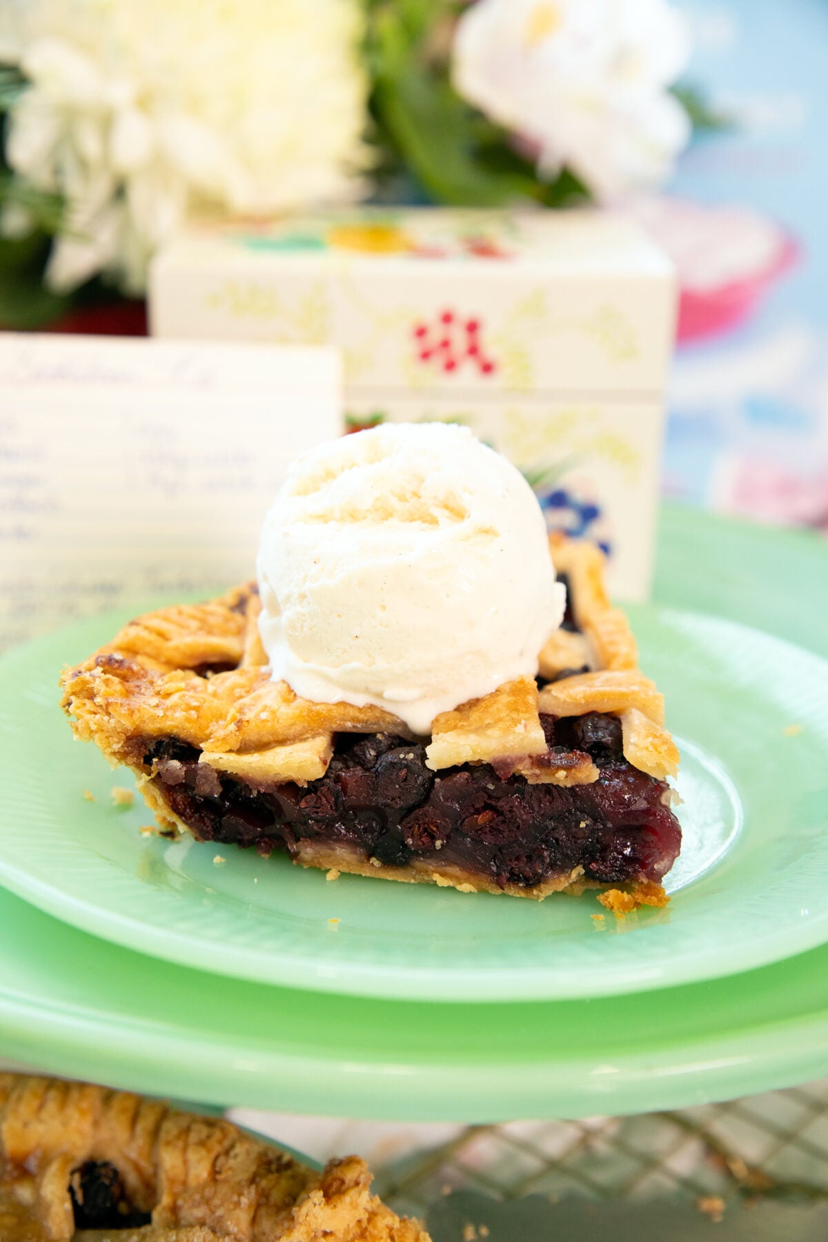 A slice of berry pie with a lattice crust, topped with a scoop of vanilla ice cream, sits on a green plate with a blurred floral background.