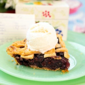 A slice of berry pie with a lattice crust, topped with a scoop of vanilla ice cream, sits on a green plate with a blurred floral background.