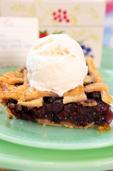 A slice of cherry pie topped with a scoop of vanilla ice cream sits on a green plate, with a handwritten recipe card blurred in the background.