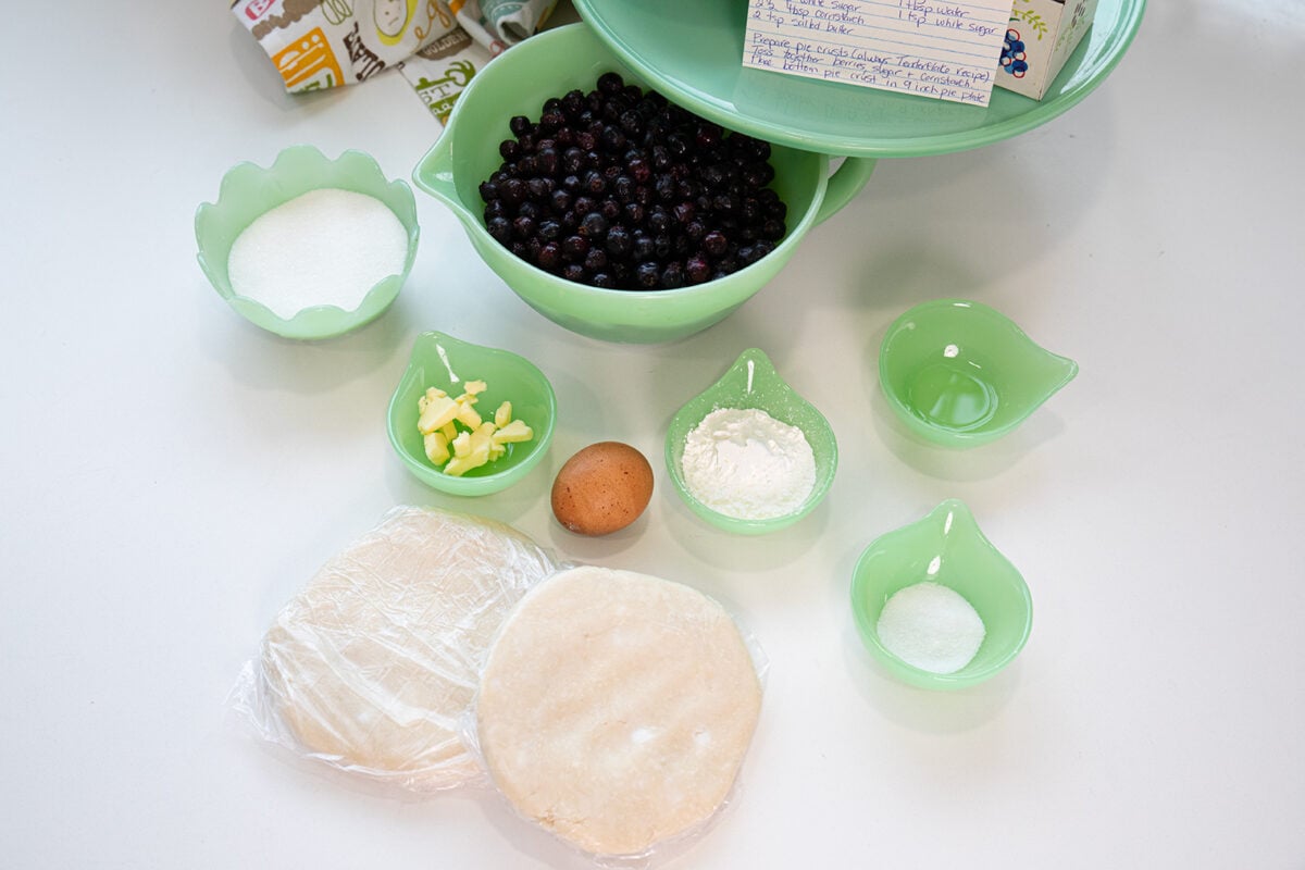 Ingredients for baking are arranged on a white surface, including blueberries, sugar, butter, an egg, flour, salt, dough, and a recipe card, all in green glass bowls.