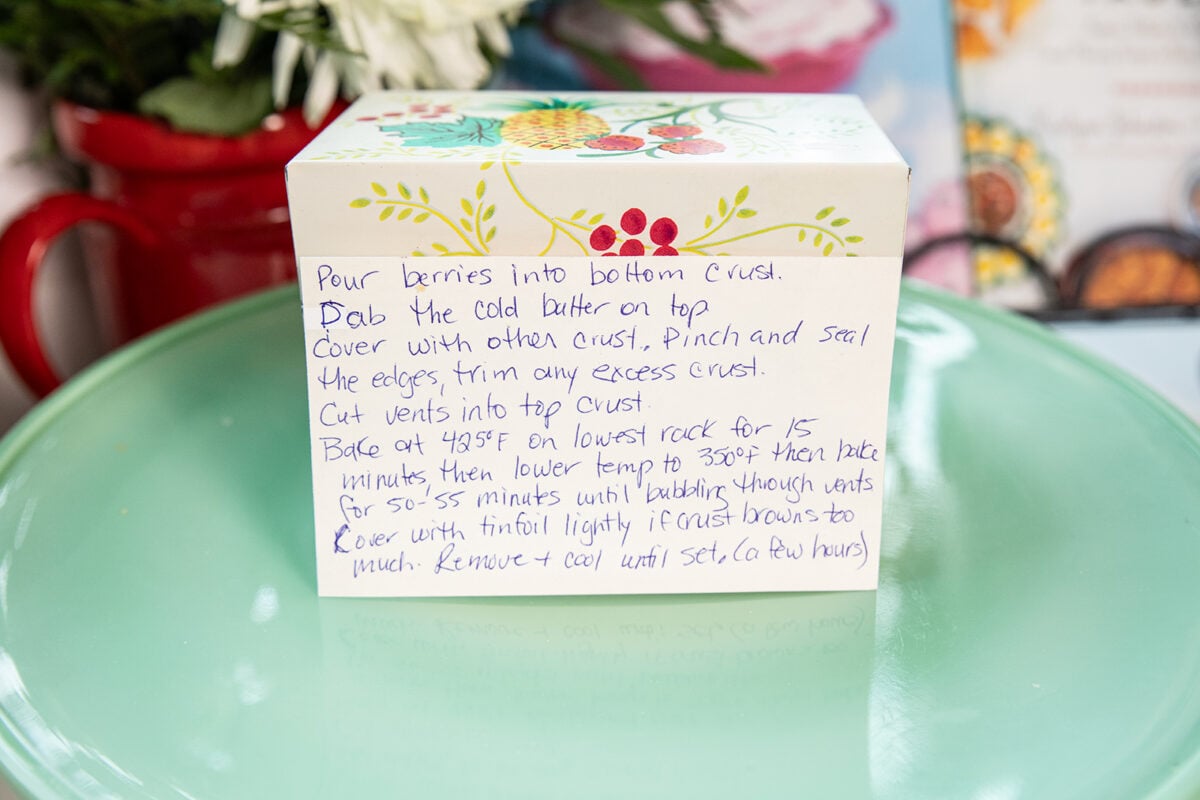 A handwritten recipe card on a floral box describes pie baking steps. The card sits on a green surface with blurred flowers in the background. The recipe mentions berries, butter, baking temperature, and cooling.