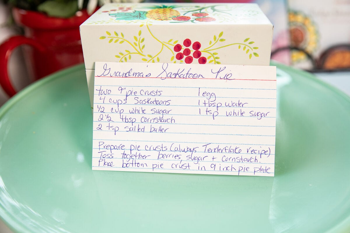 A handwritten recipe card for Grandmas Saskatoon Pie sits on a green plate with a decorative box in the background. Ingredients and instructions are written in blue ink on lined white paper.