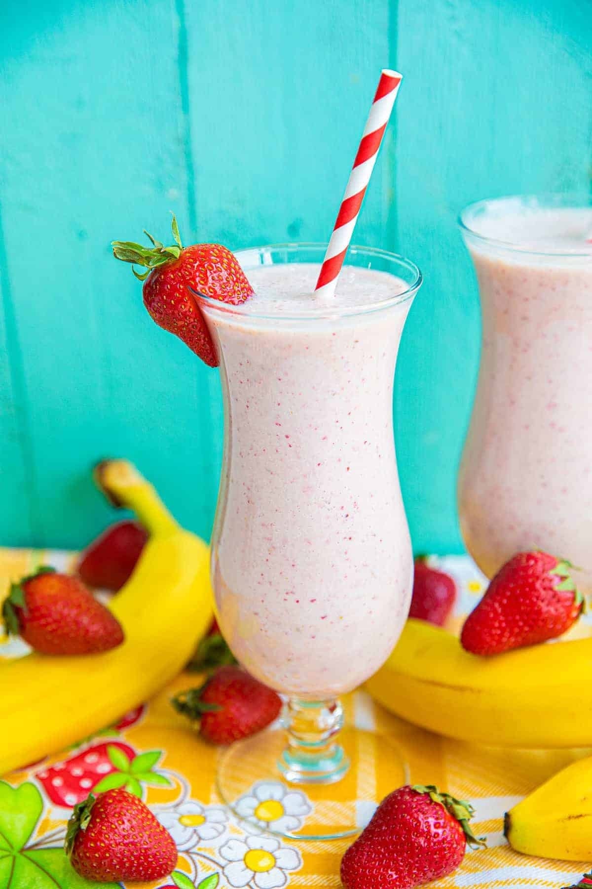 A tall glass of strawberry banana smoothie with a red and white striped straw, garnished with a strawberry. Bananas and strawberries are scattered on a colorful tablecloth, with a turquoise wooden background.