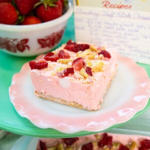 A slice of pink cheesecake topped with strawberries and nuts sits on a pastel pink and green plate. Behind it are a bowl of strawberries and a handwritten recipe card.
