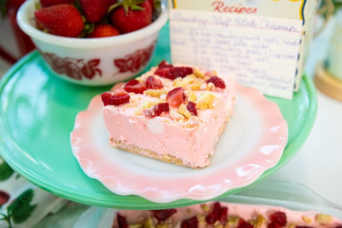 A slice of pink cheesecake topped with strawberries and nuts sits on a pastel pink and green plate. Behind it are a bowl of strawberries and a handwritten recipe card.