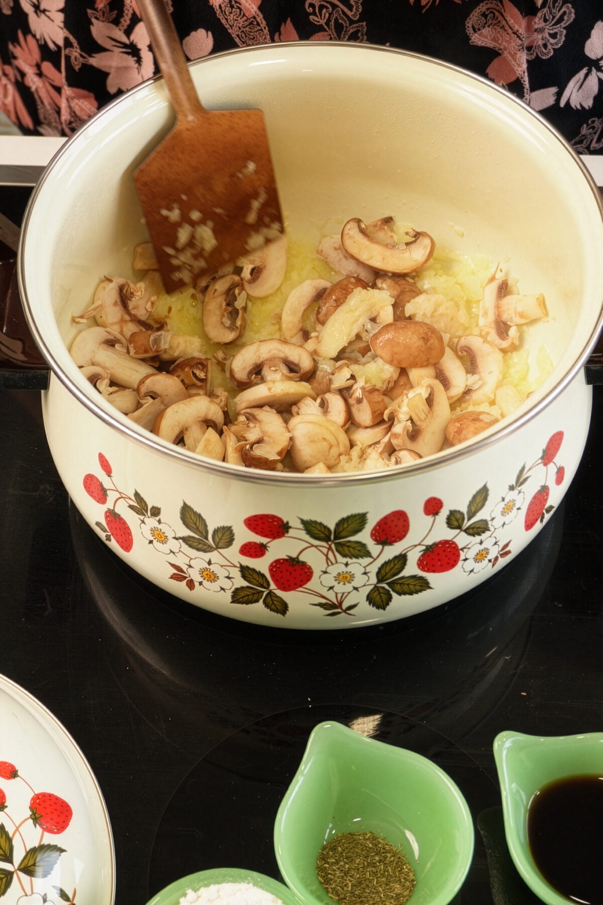 A pot with a strawberry pattern holds sliced mushrooms and onions being sautéed with a wooden spatula. Nearby are green bowls with flour, dried herbs, and a dark liquid, all on a black stovetop.
