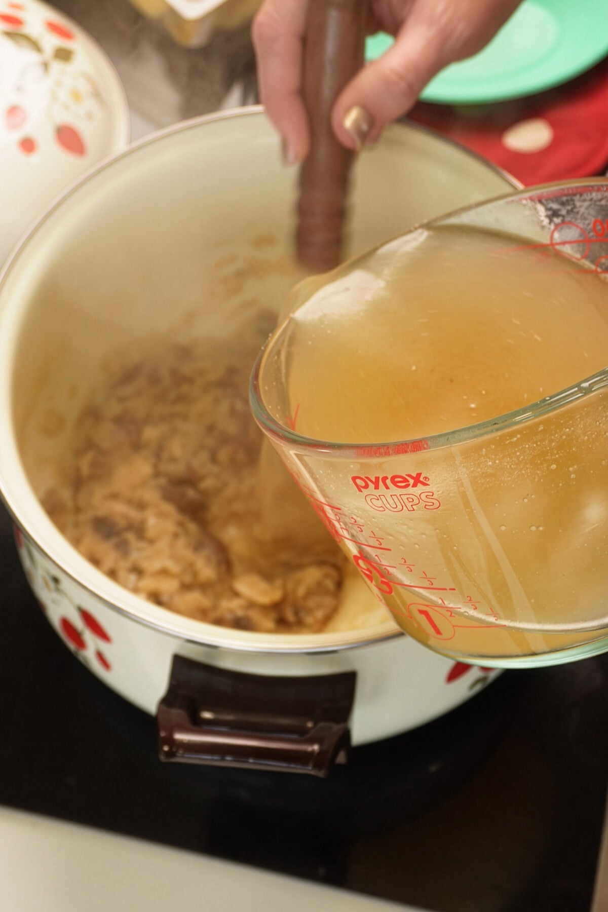 A hand pours broth from a Pyrex measuring cup into a pot with browned meat, while stirring with a wooden spoon. Steam rises from the pot on a stovetop.