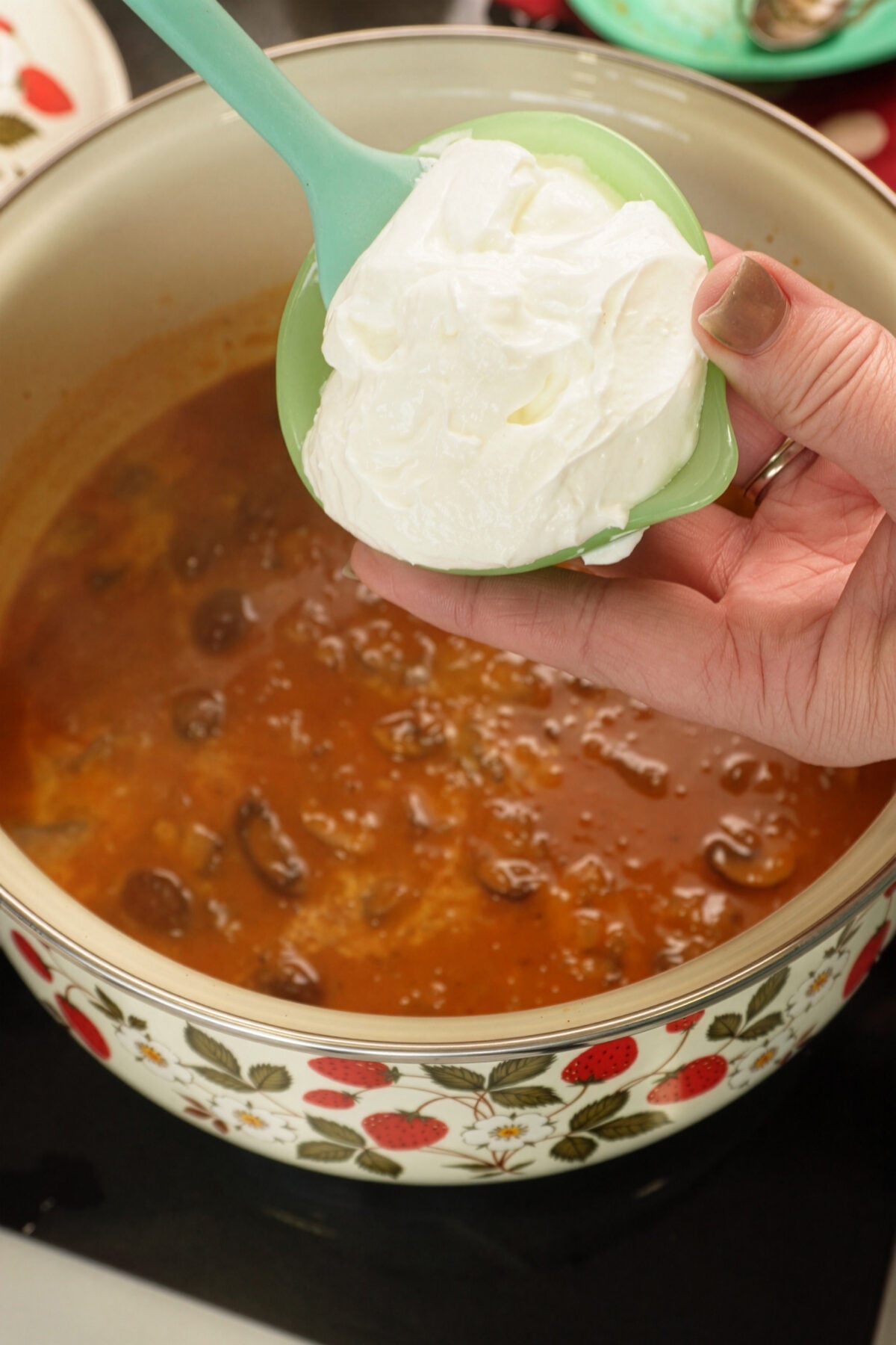 A hand holding a green bowl of sour cream above a pot of red stew with beans, ready to add the sour cream to the stew. The pot has a decorative strawberry and flower pattern.