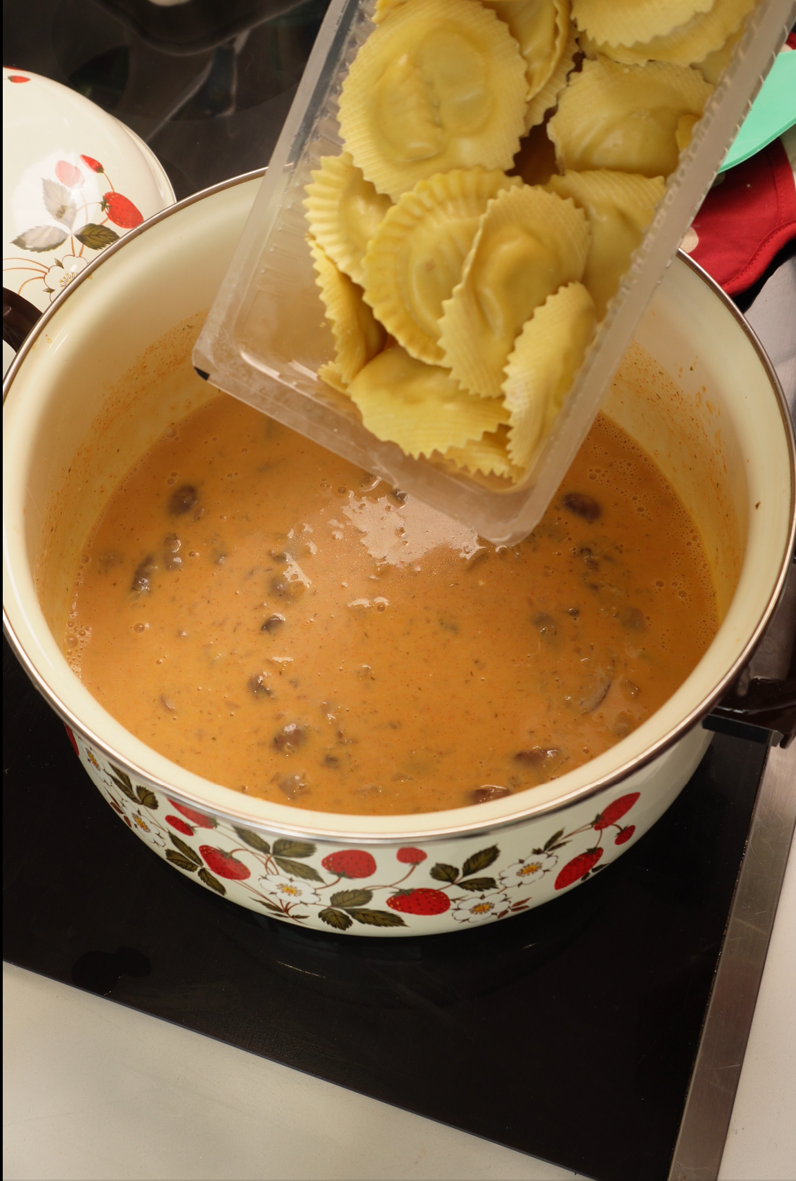 A hand pours uncooked ravioli from a plastic container into a pot of creamy tomato sauce with mushrooms, on a stove. The pot has a cherry pattern.
