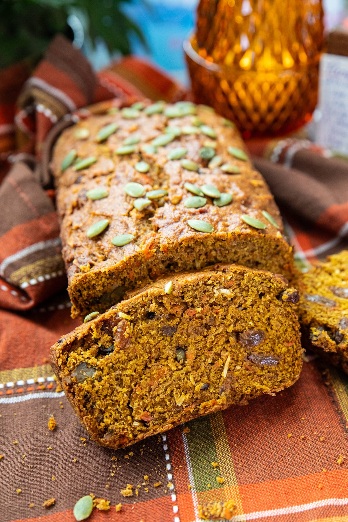 A loaf of pumpkin bread topped with pumpkin seeds, with two slices cut, sits on a plaid cloth. The bread has a moist, orange interior with visible nuts and seeds. A decorative amber glass is in the background.