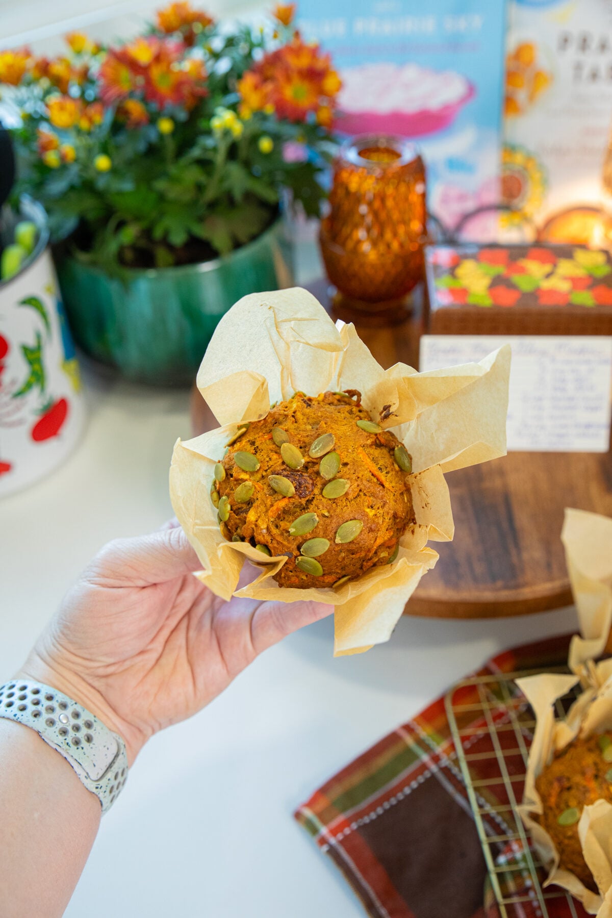 A hand holds a pumpkin muffin topped with pumpkin seeds in brown parchment paper. In the background are flowers, a candle, books, and a muffin on a cooling rack.