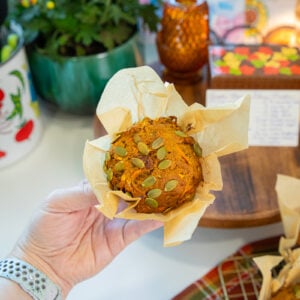 A hand holds a pumpkin muffin topped with pumpkin seeds in brown parchment paper. In the background are flowers, a candle, books, and a muffin on a cooling rack.