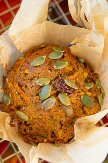 A close-up of a pumpkin muffin topped with pumpkin seeds, wrapped in parchment paper, and resting on a wire cooling rack over a colorful cloth.