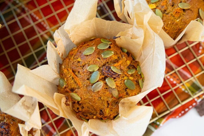 A close-up of a pumpkin muffin topped with pumpkin seeds, wrapped in parchment paper, and resting on a wire cooling rack over a colorful cloth.